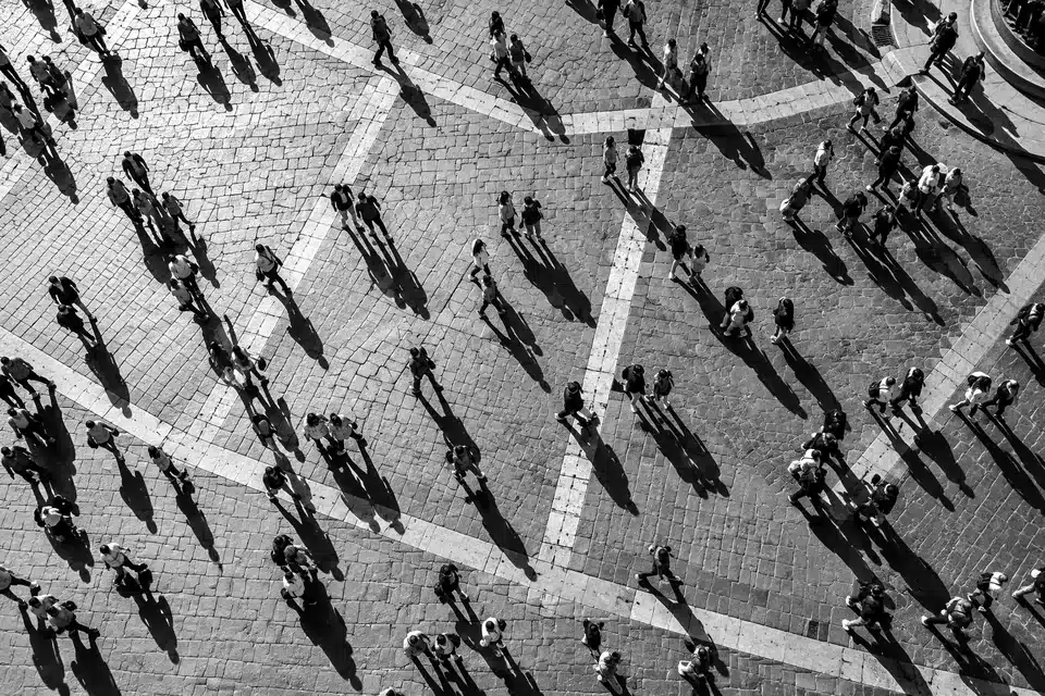 Fotografia in bianco e nero dall’alto: tante persone attraversano una piazza lastricata, con lunghe ombre.