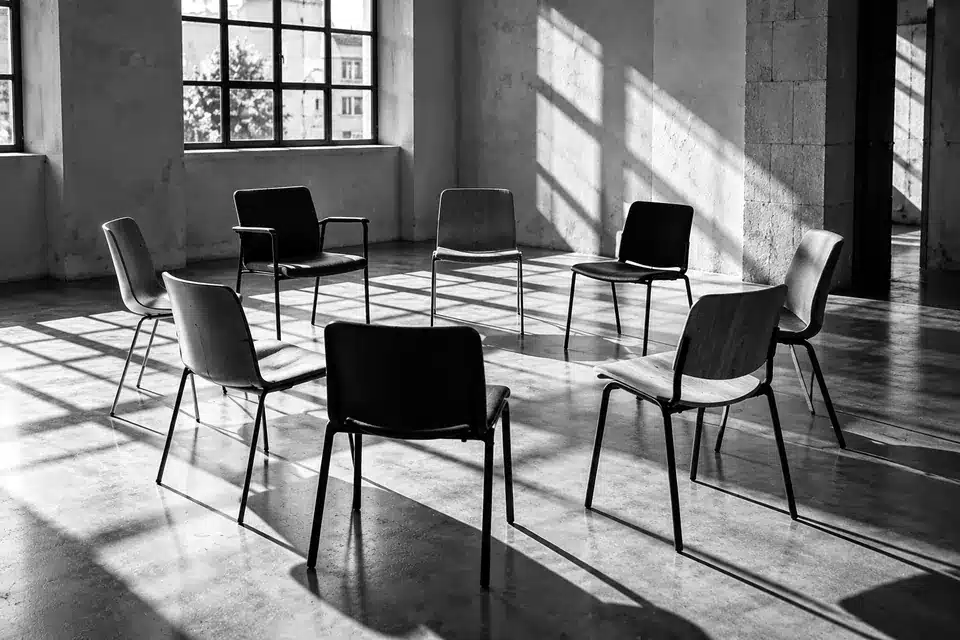 Fotografia in bianco e nero: sedie diverse disposte in cerchio in una sala luminosa, pronte per un confronto.