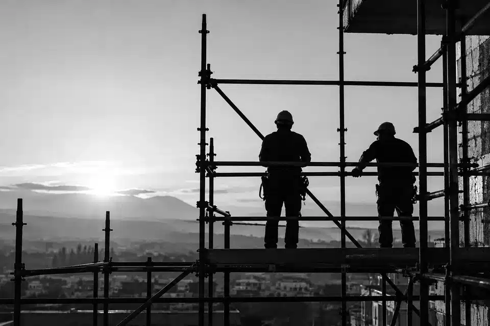 Fotografia in bianco e nero: due lavoratori con il casco, in controluce su un’impalcatura, guardano la città all’alba.